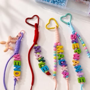 Puede incluir: Cuatro llaveros coloridos con mosquetones en forma de corazón y cordones en morado, rojo, rosa claro y azul claro. Cada llavero muestra un nombre deletreado con cuentas de letras multicolores: WENDY, MAYLE, MALYKA y STEPHANIE.