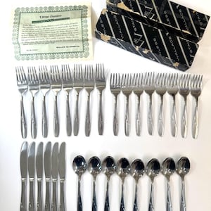May include: A Wallace stainless steel flatware set including 16 forks, 6 knives, and 8 spoons, accompanied by a black and white branded storage box and a green Lifetime Guarantee certificate with printed text detailing the product quality.