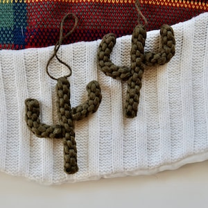 Pode incluir: Dois enfeites de cacto de crochê verde pendurados em uma superfície de malha branca. Os enfeites são feitos de fio e têm um visual rústico e feito à mão.