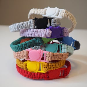 Puede incluir: Una pila de collares para gatos coloridos. Cada collar está hecho de material tejido en varios colores, incluyendo rojo, amarillo, rosa, verde y azul. Los collares tienen hebillas de plástico con un diseño de cara de gato.