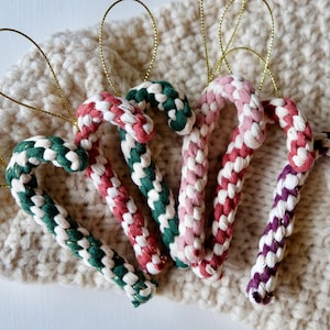 Könnte beinhalten: Fünf handgefertigte Zuckerstangen-Ornamente, jedes mit einer anderen Farbkombination. Die Ornamente sind aus weißem und farbigem Seil gefertigt und mit goldener Schnur gebunden. Die Ornamente werden auf einer cremefarbenen gestrickten Oberfläche präsentiert.