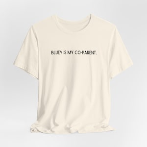 Puede incluir: Camiseta color crema con la frase "BLUEY IS MY CO-PARENT." en negro, centrada en la parte delantera. La camiseta tiene mangas cortas y cuello redondo, y está hecha de una tela suave y ligeramente arrugada.