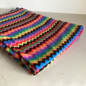 Könnte beinhalten: Gefaltete Decke mit einem lebendigen Zickzackmuster in einem Farbspektrum aus Rot, Orange, Gelb, Grün, Blau und Lila. Das Zickzack-Design ist schwarz umrandet. Die Decke scheint aus einem weichen, plüschigen Material zu sein.