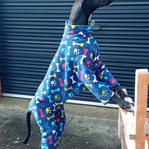 Pu&ograve; includere: Un cane bianco e nero che indossa una tuta in pile blu con un motivo a ossa e impronte di zampe. La tuta ha dettagli gialli, rossi e blu chiaro.