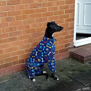 Pu&ograve; includere: Un cane nero che indossa un cappotto in pile blu per cani con un motivo a ossa e impronte di zampe. Il cane &egrave; seduto su una superficie di cemento grigia davanti a un muro di mattoni.