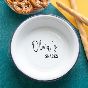 May include: White ceramic bowl with a black rim, featuring the text "Olivia's Snacks" on a teal background.