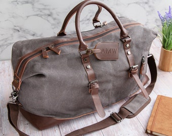 Pode incluir: Uma bolsa de viagem de lona cinza e couro marrom com uma etiqueta de bagagem de couro que diz "AMW". A bolsa tem uma alça superior, uma alça de ombro e duas fivelas laterais.