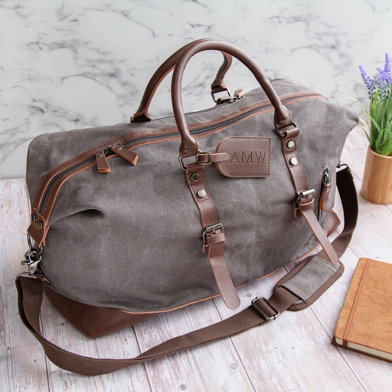 May include: A gray canvas and brown leather duffel bag with a leather luggage tag that says "AMW". The bag has a top handle, a shoulder strap, and two side buckles.