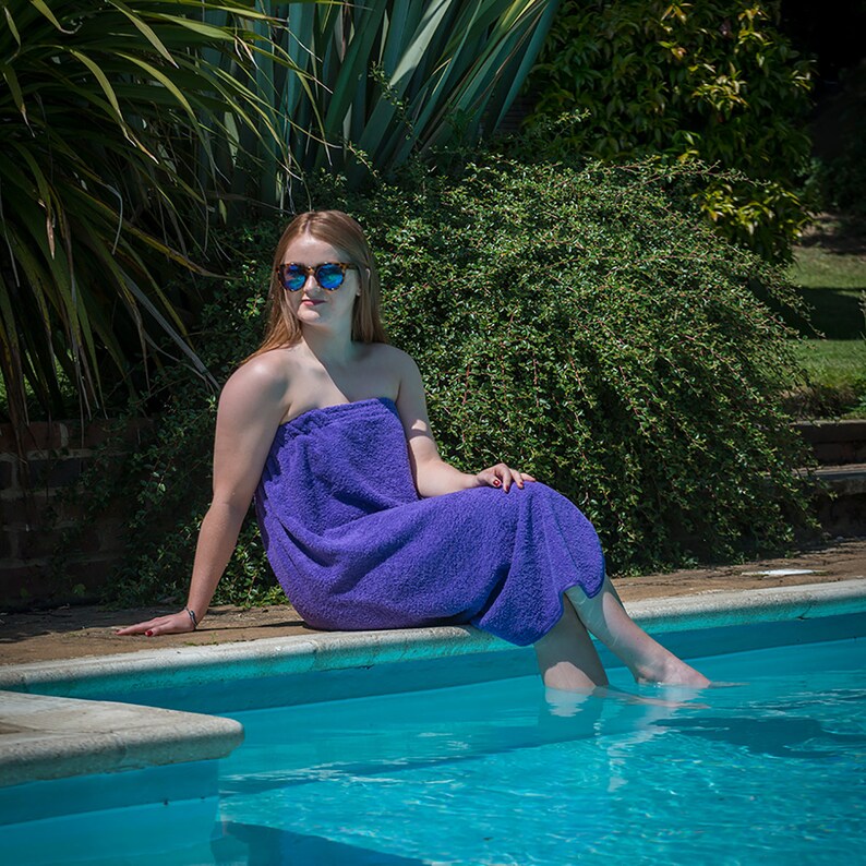 May include: A woman wearing sunglasses sits on the edge of a swimming pool with her feet in the water. She is wearing a purple towel wrapped around her.