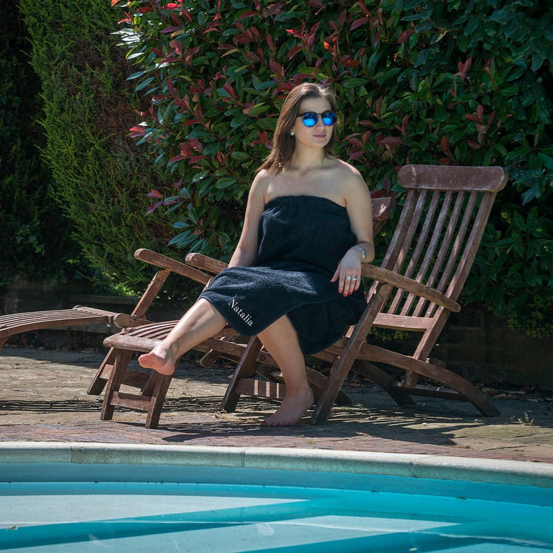 May include: A woman wearing sunglasses sits on a wooden lounge chair by a swimming pool. She is wearing a black towel with the name "Natalia" embroidered on it.