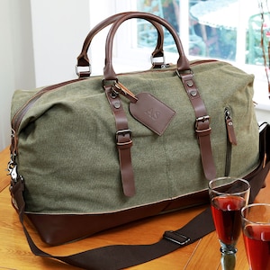 May include: A green canvas duffel bag with brown leather accents and a brown leather luggage tag with the initials "AS" embossed on it. The bag has a long adjustable shoulder strap and two top handles.