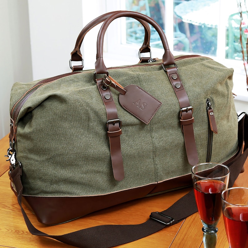 May include: A green canvas duffel bag with brown leather accents and a brown leather luggage tag with the initials "AS" embossed on it. The bag has a long adjustable shoulder strap and two top handles.