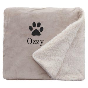 May include: A beige and white sherpa blanket with a black paw print and the name "OzZy" embroidered on it.