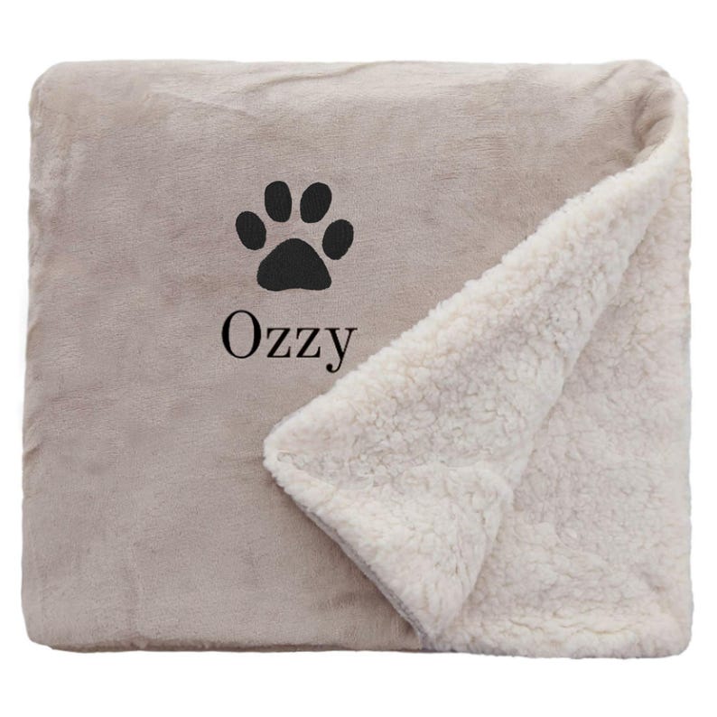 May include: A beige and white sherpa blanket with a black paw print and the name "OzZy" embroidered on it.