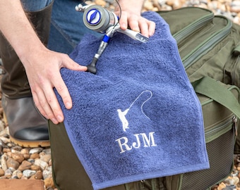 May include: A navy blue rectangular cotton towel featuring an embroidered white graphic of a person fishing with a rod and the letters RJM underneath. The towel is held over a dark green fishing bag next to a silver and blue fishing reel.