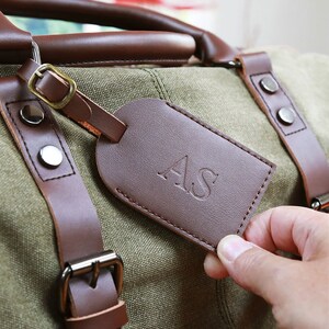 May include: A brown leather luggage tag with the initials "AS" embossed in the center. The tag is attached to a green canvas duffel bag with brown leather straps.