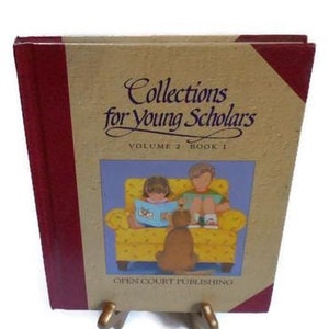 Może przedstawiać: Brązowa książka z czerwoną okładką zatytułowana "Collections for Young Scholars, Volume 2, Book 1". Okładka przedstawia ilustrację dwojga dzieci czytających w żółtym fotelu z psem.