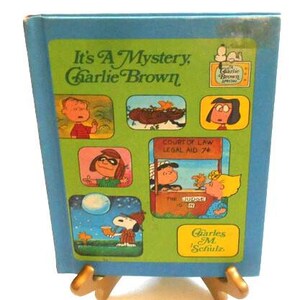 May include: A blue and green paperback book titled "It's A Mystery. Charlie Brown."  The cover features illustrations of the Peanuts characters, including Charlie Brown, Snoopy, Lucy, and Linus. The book is on a gold metal stand.