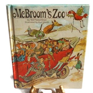 Puede incluir: Un libro de tapa dura titulado "McBroom's Zoo" de Sid Fleischman, ilustrado por Kurt Werth. La portada presenta una colorida ilustración de un vehículo rojo lleno de gente, con una escena animalística caprichosa en el fondo. El libro se exhibe sobre un soporte dorado.