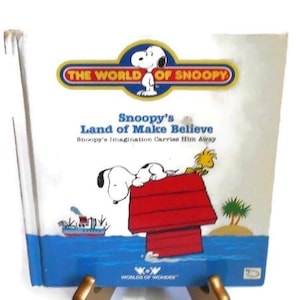 May include: A vintage children's book titled "Snoopy's Land of Make Believe" is displayed on a gold-colored stand. The cover features Snoopy on his red doghouse, with a blue ocean scene and the words "The World of Snoopy".