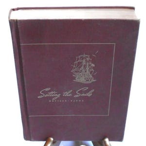 Pode incluir: Um livro castanho-avermelhado intitulado "Setting the Sails" com uma ilustração dourada de um veleiro. O livro está apoiado num suporte dourado sobre um fundo branco. A capa tem um design retangular.