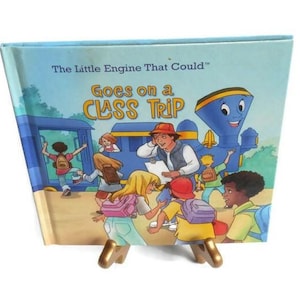 Pode incluir: Um livro infantil azul e amarelo intitulado "The Little Engine That Could™ Goes on a Class Trip". A capa apresenta uma locomotiva de desenho animado com um rosto sorridente e um grupo de crianças entrando no trem.