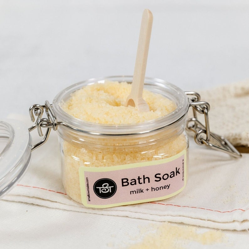 Puede incluir: Un tarro de vidrio transparente con tapa plateada y una cuchara de madera en su interior. El tarro est&aacute; lleno de un ba&ntilde;o de inmersi&oacute;n amarillo y tiene una etiqueta que dice "Bath Soak milk + honey".