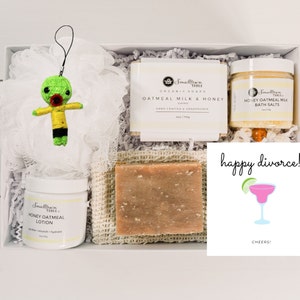 Puede incluir: Una caja de regalo blanca llena de productos para el ba&ntilde;o y el cuerpo. La caja contiene un llavero de mu&ntilde;eca vud&uacute; verde, una botella blanca de loci&oacute;n de avena y miel, una barra de jab&oacute;n de leche de avena y miel, un tarro de sales de ba&ntilde;o de leche de avena y miel y una tarjeta que dice "&iexcl;feliz divorcio!" con una ilustraci&oacute;n de margarita.