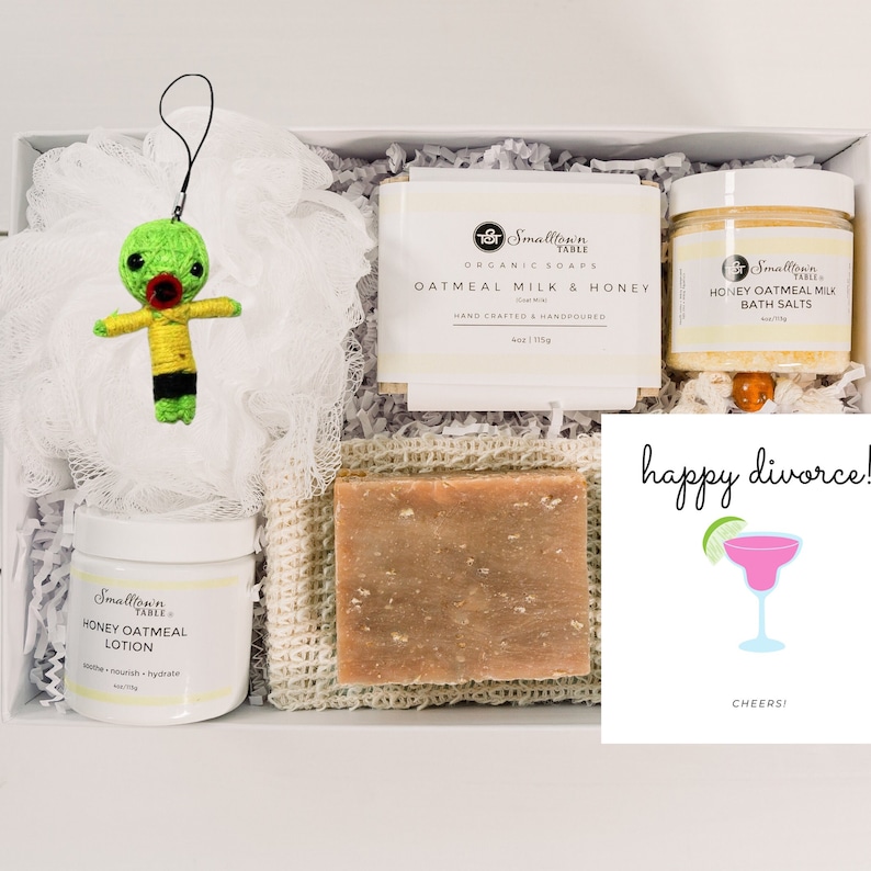Puede incluir: Una caja de regalo blanca llena de productos para el ba&ntilde;o y el cuerpo. La caja contiene un llavero de mu&ntilde;eca vud&uacute; verde, una botella blanca de loci&oacute;n de avena y miel, una barra de jab&oacute;n de leche de avena y miel, un tarro de sales de ba&ntilde;o de leche de avena y miel y una tarjeta que dice "&iexcl;feliz divorcio!" con una ilustraci&oacute;n de margarita.