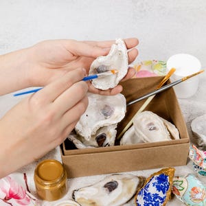 Puede incluir: Una persona pinta el interior de una concha de ostra con un pequeño pincel azul. La concha de ostra es una de las muchas que hay en una caja de cartón. Hay dos pequeños frascos de pintura dorada y un frasco de pintura blanca sobre la mesa.