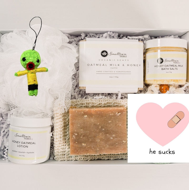 Puede incluir: Una caja de regalo blanca llena de productos para el ba&ntilde;o y el cuerpo. La caja contiene un llavero de mu&ntilde;eca vud&uacute; verde, un frasco blanco de loci&oacute;n de miel y avena, una barra de jab&oacute;n de leche de avena y miel, una barra de jab&oacute;n y un frasco de sales de ba&ntilde;o de leche de avena y miel. La caja tambi&eacute;n incluye una tarjeta rosa con un coraz&oacute;n y una tirita y el texto "apesta".