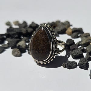 May include: A teardrop-shaped ring featuring a dark brown, speckled stone set in a silver frame. The setting is decorated with a beaded edge and a twisted rope detail. The band of the ring is split into two thin silver wires that meet at the back.