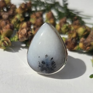 May include: A teardrop-shaped ring featuring a smooth, milky white stone with dark, fern-like dendritic inclusions at the base. The gemstone is set in a polished silver metal bezel, creating a clean, reflective border around the organic pattern.
