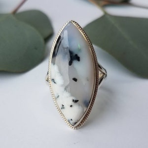 May include: A silver ring with a large, oval-shaped stone. The stone is white with black and gray inclusions, set in a silver band with a textured edge. The ring is displayed on a white surface with green leaves in the background.
