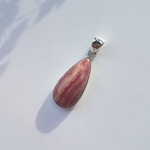 May include: A teardrop-shaped pendant with a pink and white stone set in a silver-colored metal frame. The stone has a layered appearance with varying shades of pink and white. The pendant is displayed on a white surface.