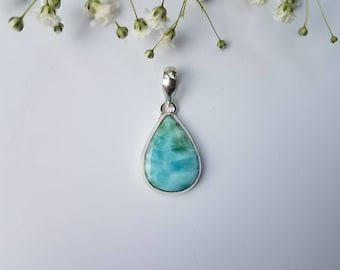 May include: A teardrop-shaped pendant with a light blue and green stone set in a silver bezel. The pendant hangs from a silver bail. White flowers are in the background.