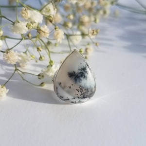 May include: A silver ring featuring a large, teardrop-shaped dendritic agate stone. The stone has a white base with black and gray fern-like inclusions. Delicate white flowers are in the background, creating a soft, natural aesthetic.