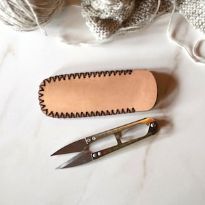 May include: A light brown leather needle case with black stitching and a pair of silver thread snips. The case is rectangular with rounded ends. The thread snips are open, ready for use.