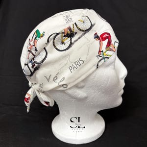 May include: White cycling-themed scrub cap featuring illustrations of cyclists, bicycles, and the word "Paris." The cap has a tie closure and is displayed on a mannequin head. The design includes a map-like layout.