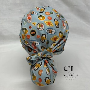 May include: A light blue scrub cap with a repeating pattern of fruit-themed stickers. The stickers feature images of bananas, papayas, and other fruits, with text that reads "Sun Ripe" and "Papaya". The cap is tied in a bow at the back.