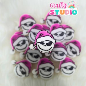 May include: A collection of pink and white cookies decorated with the face of Jack Skellington from the Nightmare Before Christmas movie. The cookies are shaped like a head with a pink Santa hat on top. The cookies are arranged in a circle on a white surface. The text "crafty STUDIO" is visible in the background.