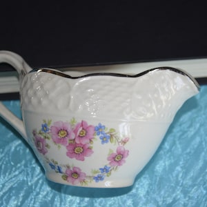 May include: White ceramic creamer with a scalloped edge and a floral design featuring pink and blue flowers. The creamer has a silver trim around the top edge.