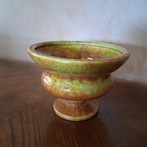 May include: A ceramic pedestal bowl with a wide, shallow top and a rounded base. The bowl features a speckled brown and green glaze, with a lighter green rim. The bowl is likely vintage and suitable for decorative purposes.
