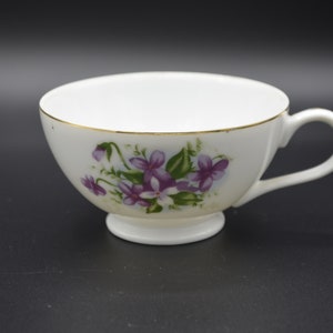 May include: A white teacup with a gold rim and a floral design featuring purple flowers and green leaves.