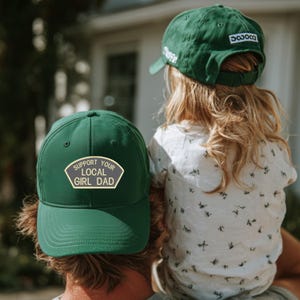 Könnte beinhalten: Zwei grüne Baseballkappen. Eine Kappe hat den Text "SUPPORT YOUR LOCAL GIRL DAD" auf einem Aufnäher. Die andere Kappe hat den Text "savoca". Die Kappen werden von einer Person und einem Kind getragen.