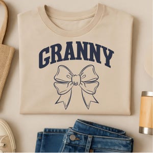 May include: A beige t-shirt with the word "GRANNY" in navy blue arched above a navy blue bow graphic. The shirt is folded and placed on a flat surface with blue jeans, a tote bag, and a shoe.