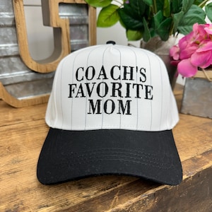 May include: A white baseball cap with black pinstripes and a black brim. The text "COACH'S FAVORITE MOM" is embroidered in black on the front of the cap. The cap is sitting on a wooden surface.
