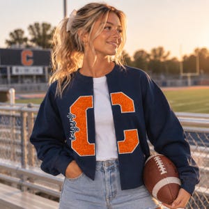 May include: A navy blue cropped sweatshirt featuring a white crew neck shirt underneath. The sweatshirt has large, glittery orange block letters on the front, with the word "cougars" written in white script along the side of the left letter.