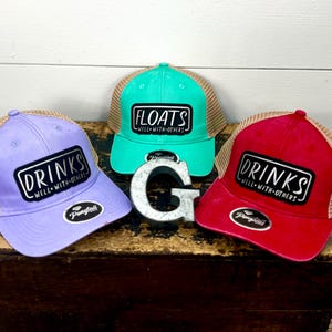 May include: Three trucker hats with mesh back panels. The hats are in different colors: purple, teal, and red. Each hat has a black patch with white text that reads "Drinks Well With Others" or "Floats Well With Others".