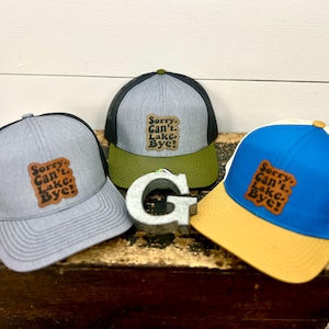 May include: Three trucker hats with a brown leather patch that says "Sorry. Can't. Lake. Bye!" The hats are gray, blue, and green.